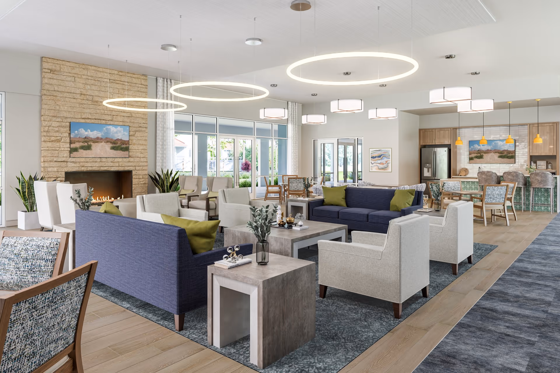 A spacious and modern senior living common area featuring comfortable seating with blue and beige sofas and armchairs arranged around coffee tables. The room has large windows allowing natural light, a stone fireplace with a landscape painting above it, and contemporary circular and square ceiling lights. In the background, there is a dining area with tables and chairs, a kitchen area with a refrigerator, and pendant lights hanging over a counter with bar stools.