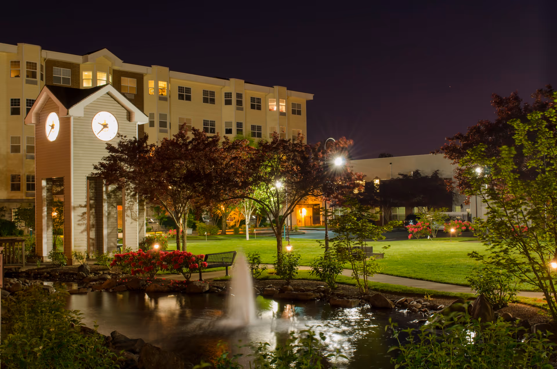 Night view of Cherrywood Village a Generations Community showing a well-lit outdoor garden area with a pond and fountain in the foreground, a clock tower, trees, benches, and a multi-story building in the background.