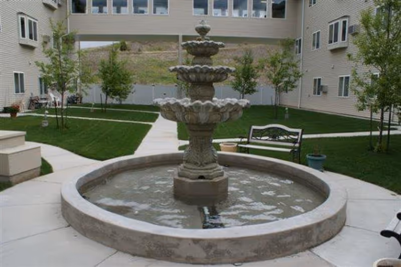 Stone three-tier fountain in a courtyard with benches, walkways, grass and surrounding apartment buildings.