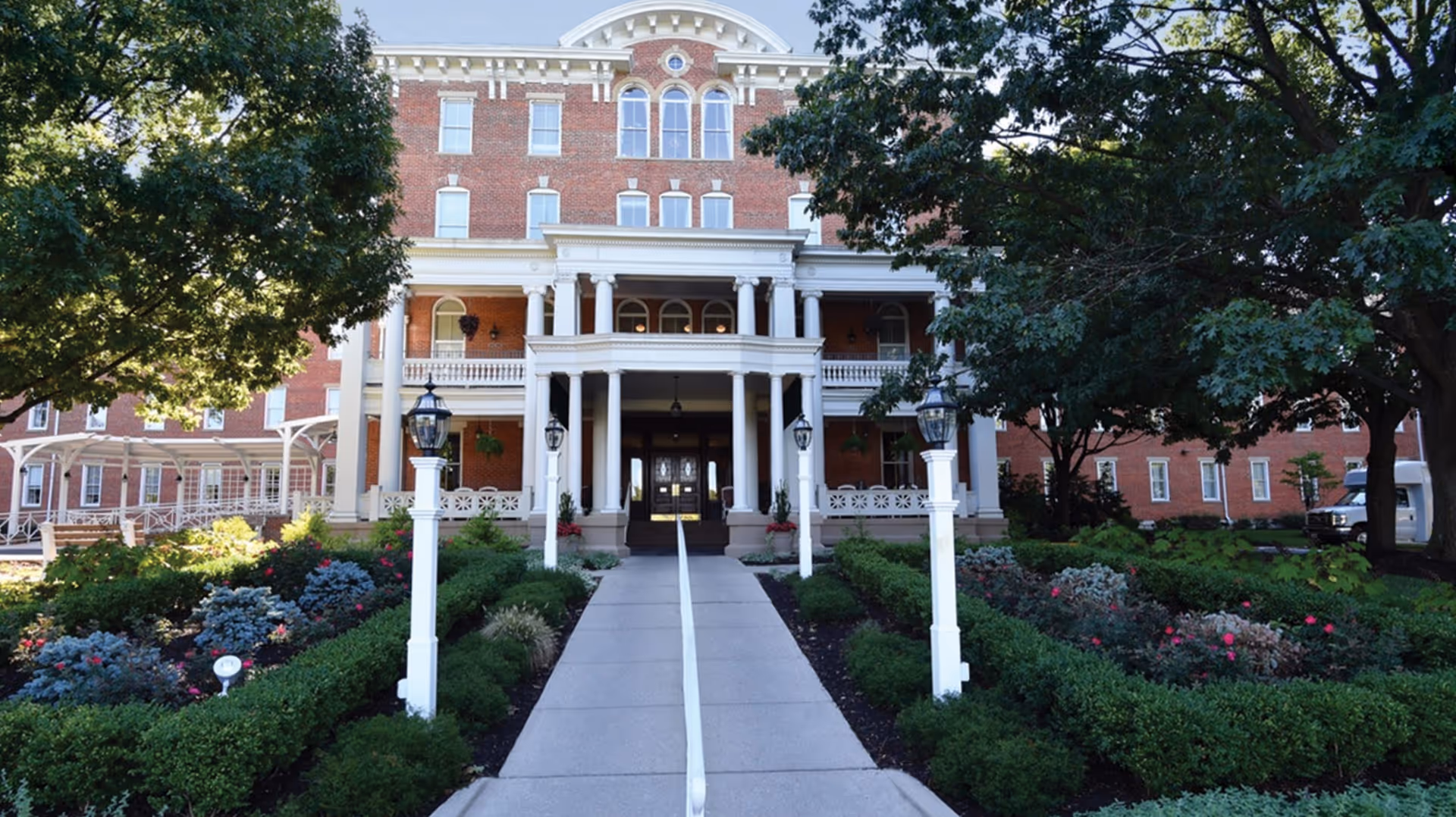 Front exterior view of a large brick retirement community building with white columns and a covered entrance. The pathway leading to the entrance is flanked by white lamp posts and well-maintained gardens with shrubs and flowers. Trees provide shade on either side of the building.