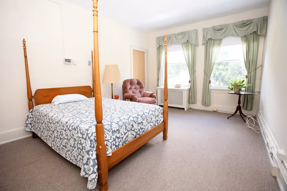 A bright bedroom with a wooden four-poster bed covered with a patterned blue and white bedspread, a cushioned armchair, a floor lamp, two large windows with green curtains, a small round table with a plant, and beige carpeting.