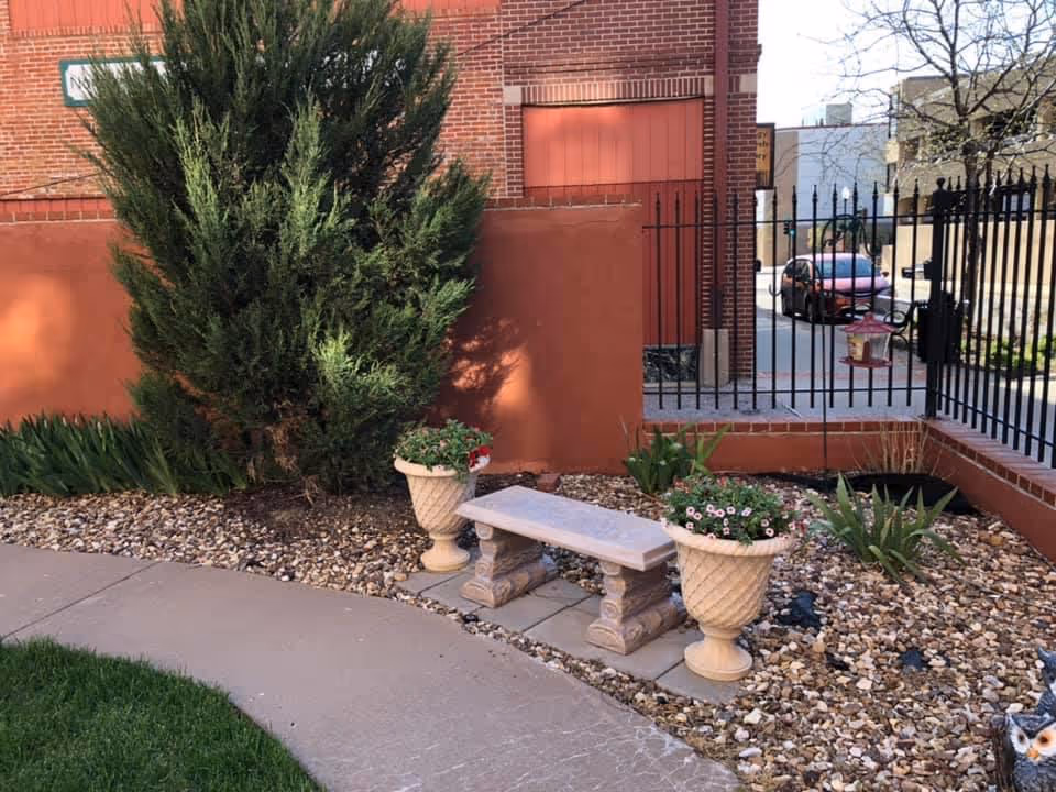 Outdoor garden area with a stone bench flanked by two large decorative planters filled with flowers. The area is landscaped with rocks and plants, bordered by a curved concrete pathway and a grassy lawn. In the background, there is a red brick building, a black metal fence, and a parked car visible through the fence.