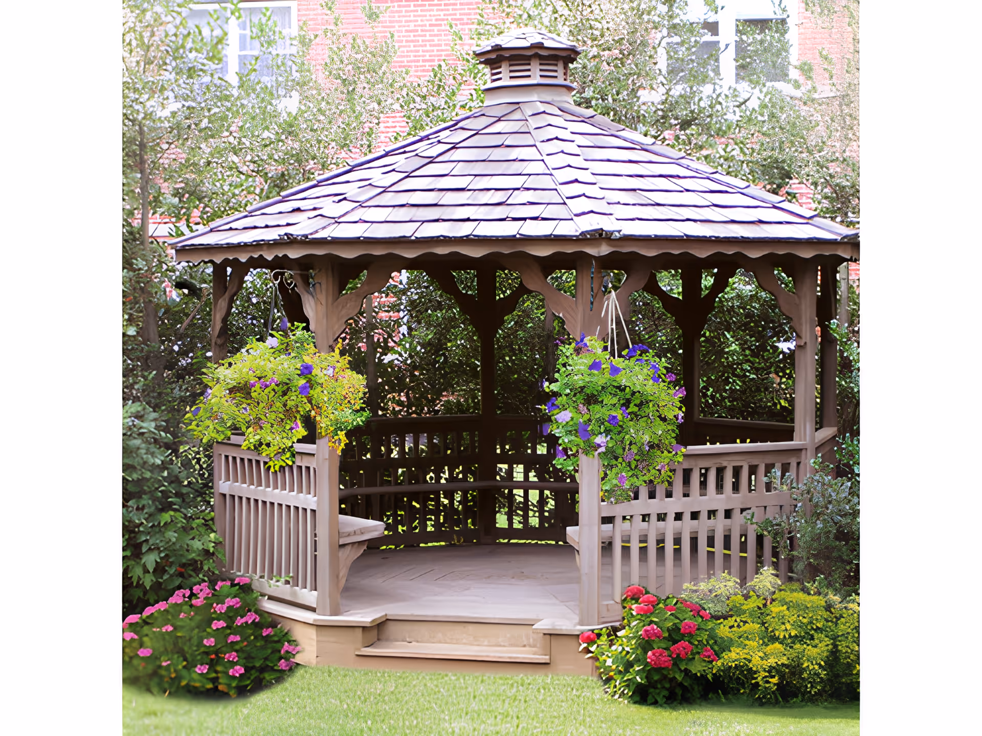 A wooden gazebo with a shingled roof surrounded by lush greenery and colorful flowers, including hanging baskets with purple and yellow flowers, located in a garden area with a brick building partially visible in the background.