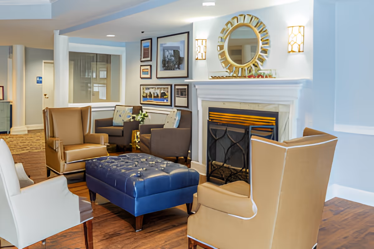 A cozy living room area with a white fireplace, a round decorative mirror above it, and two wall sconces on either side. The room features a blue tufted ottoman in the center surrounded by beige and white armchairs. There are additional seating areas with dark brown chairs and framed artwork on the walls. The floor is wooden, and the overall ambiance is warm and inviting.