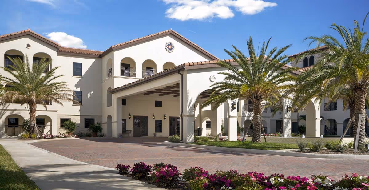 Exterior view of a senior living facility with a Mediterranean architectural style featuring white stucco walls, arched windows and doorways, and a tiled roof. The entrance has a covered porte-cochère supported by columns. Palm trees and colorful flowers line the driveway and landscaping under a blue sky with some clouds.