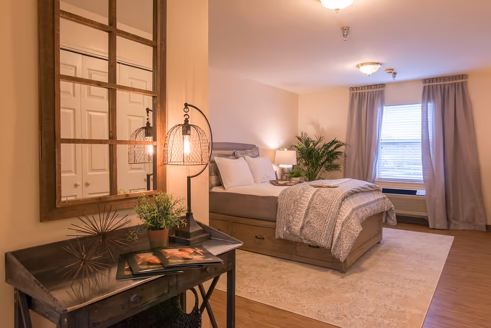 A cozy bedroom with a neatly made bed featuring white pillows and a patterned comforter. A wooden nightstand with a lamp and a green plant is beside the bed. A large window with curtains allows natural light into the room. In the foreground, there is a small table with a decorative lamp, a potted plant, and some magazines, with a large wooden-framed mirror hanging above it.
