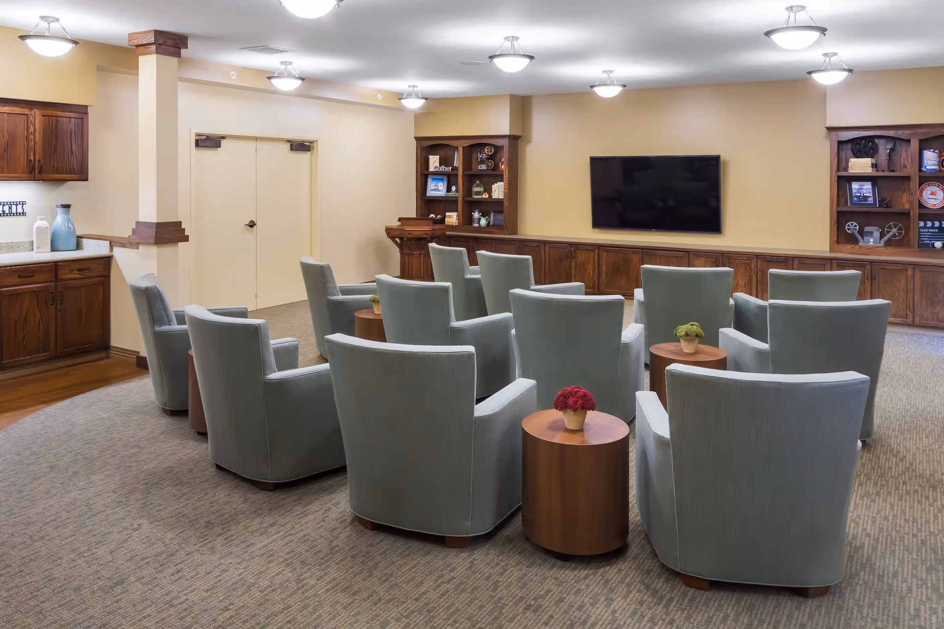 A cozy common area with multiple light blue armchairs arranged in rows facing a wall-mounted flat screen TV. The room features beige walls, carpeted flooring, wooden cabinetry along the back wall with decorative items, and small round wooden tables with potted plants between some chairs. Ceiling lights provide bright illumination.