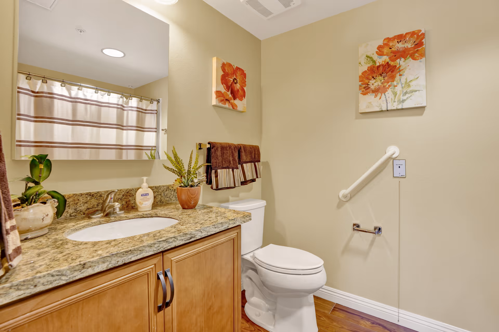 A clean and well-lit bathroom featuring a granite countertop with a sink, a large mirror, a toilet with a safety grab bar on the wall beside it, and decorative orange floral paintings on the beige walls. There are brown and beige striped towels hanging on a towel rack and a shower curtain with similar stripes visible in the mirror reflection.