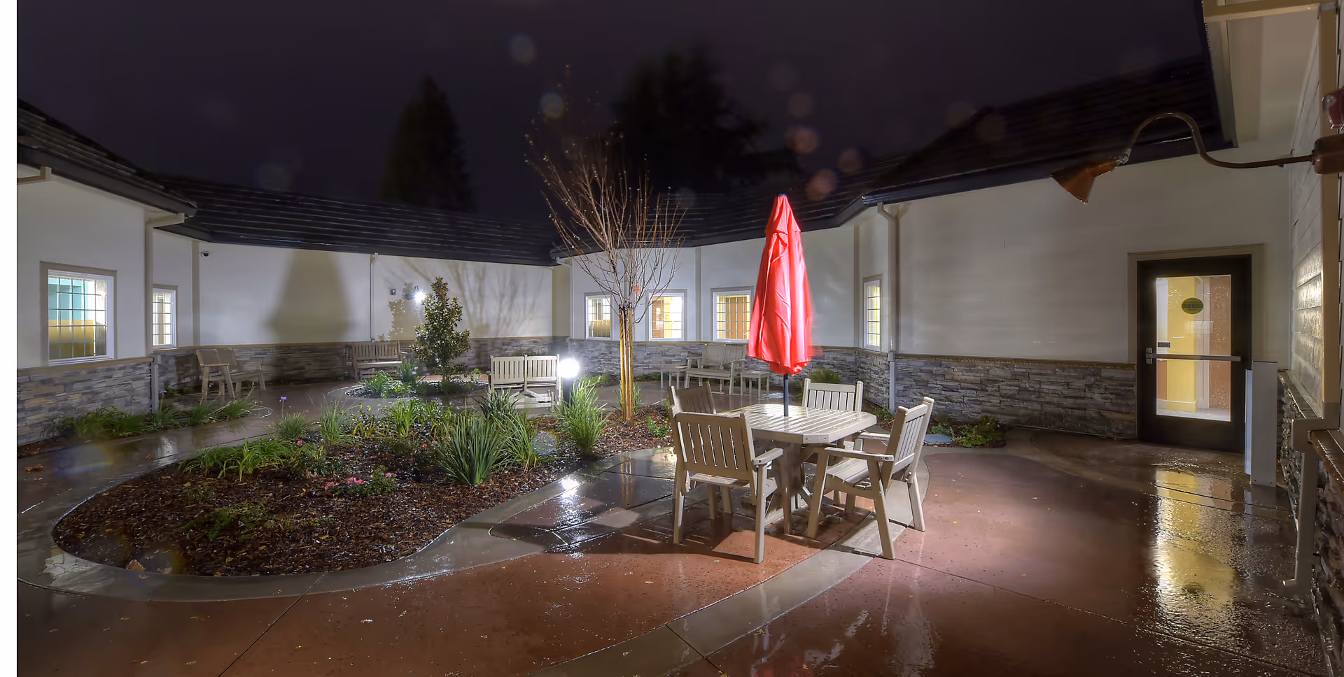 Night view of a courtyard at a senior living community with a round garden bed in the center, surrounded by a wet concrete patio. There is a table with four chairs and a closed red umbrella. The courtyard is enclosed by a building with lit windows and a door.