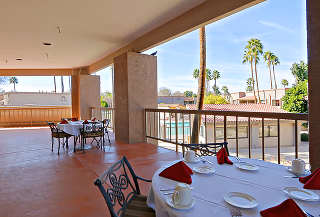 Covered outdoor patio area with tables set for dining, featuring white tablecloths, red napkins, white plates, and coffee cups. The patio overlooks a pool area and is surrounded by palm trees and other buildings under a clear blue sky.