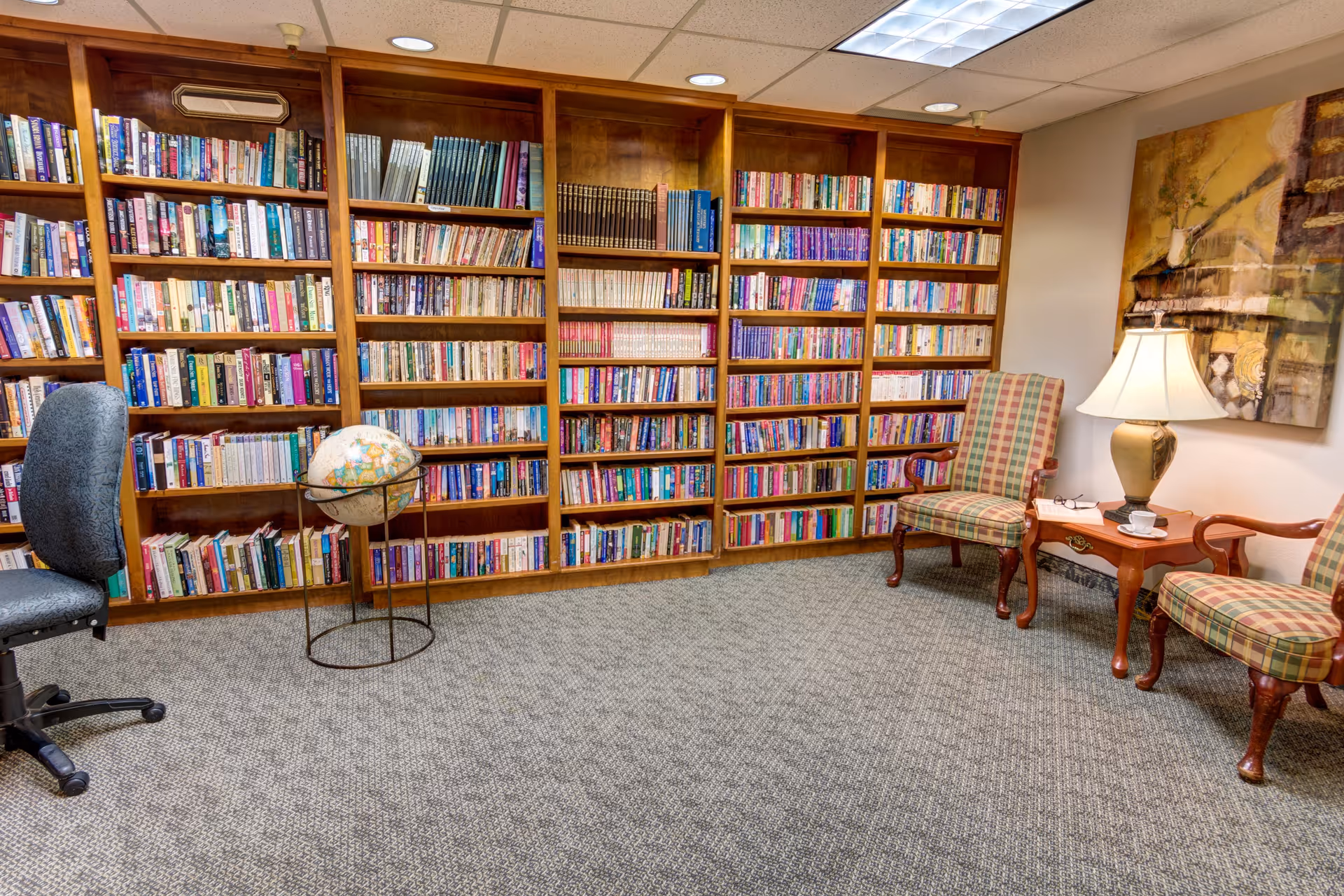 A cozy library room with wooden bookshelves filled with books along the wall. There is a globe on a stand, two plaid upholstered chairs, a wooden side table with a lamp, and a framed painting on the wall. A gray office chair is partially visible on the left side.