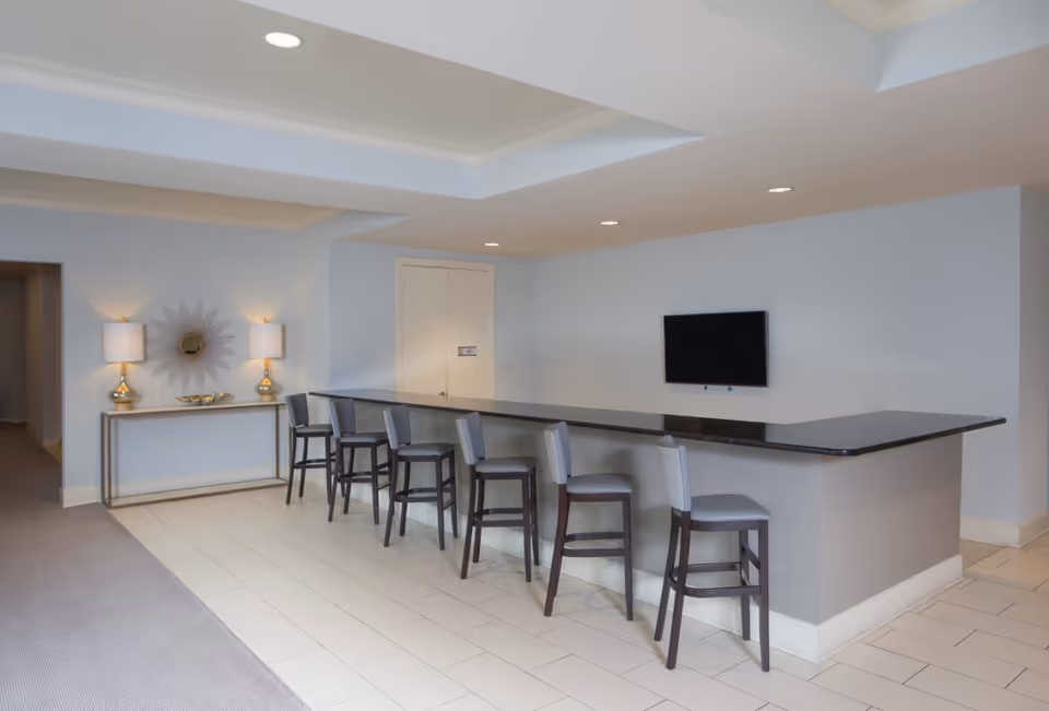 Communal lounge area with a long bar counter, five bar stools, a wall-mounted TV and a console table with lamps and decorative mirror.