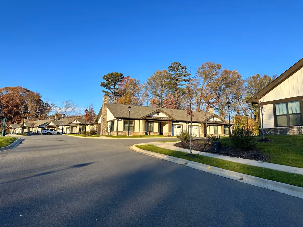 Row of single-story residential buildings with garages along a curved paved road and landscaped lawns under a clear blue sky.
