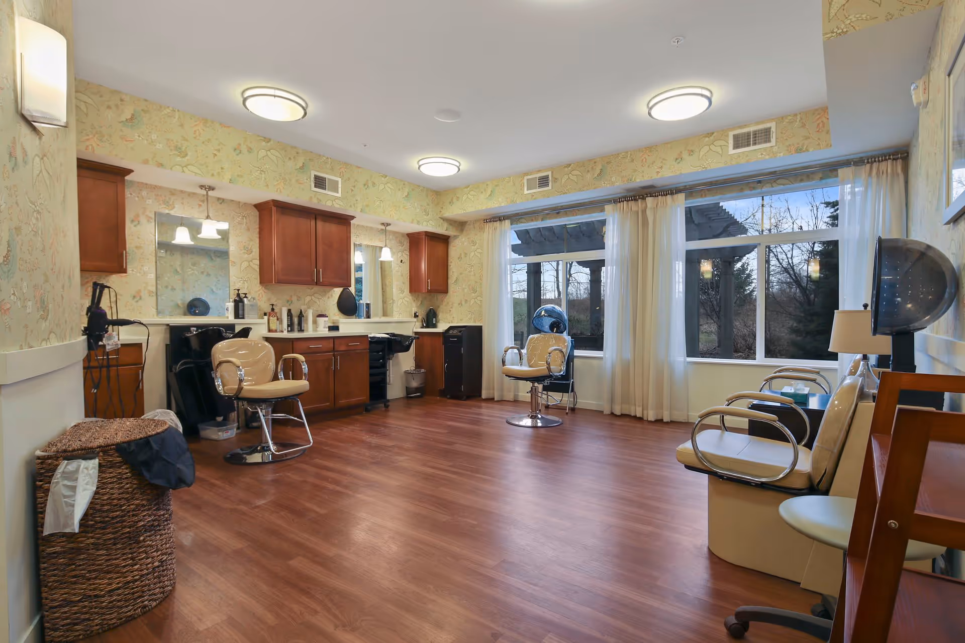 A spacious hair salon room with wooden flooring, two salon chairs, large windows with sheer curtains letting in natural light, wooden cabinets with mirrors and hair care products, and a hair dryer chair on the right side.