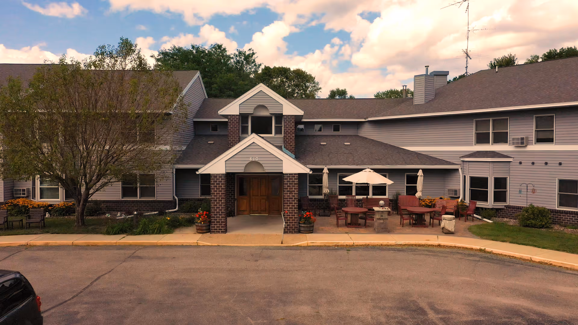 Exterior view of a two-story assisted living facility building with a covered entrance supported by brick columns. There is a patio area with tables, chairs, and umbrellas to the right of the entrance. Trees and shrubs surround the building under a partly cloudy sky.