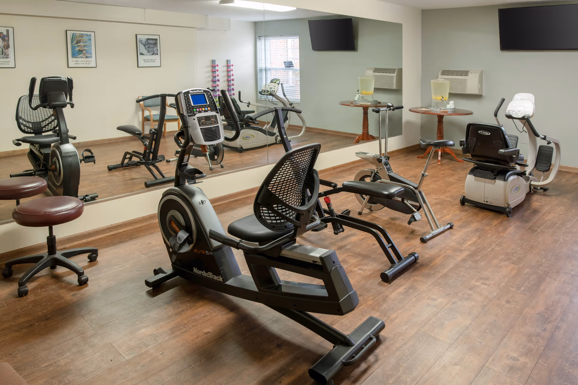 A small fitness room with exercise equipment including a recumbent bike, a rowing machine, and a stationary bike. The room has wooden flooring, a large mirror on one wall, two small round tables with pitchers of water and cups, a wall-mounted TV, and framed pictures on the wall.