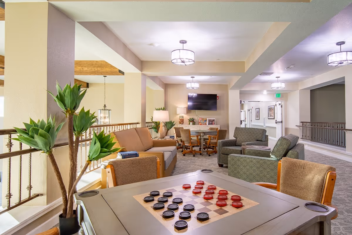A bright and spacious senior living common area with comfortable seating including sofas and armchairs arranged around a checkers game table. The room features modern light fixtures, potted plants, a wall-mounted TV, and a table with chairs in the background. The space has a warm and inviting atmosphere with beige walls and carpeted floors.