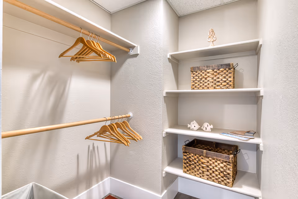 A small walk-in closet with two wooden hanging rods holding wooden hangers on the left side. On the right side, there are white built-in shelves holding two woven baskets, two small decorative fish figurines, and some brochures or papers. The walls are light-colored and the floor is dark wood.