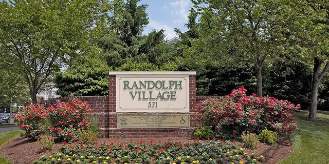 A brick and stone sign for Randolph Village with the number 531, surrounded by well-maintained landscaping including green bushes, red and yellow flowers, and trees in the background under a partly cloudy sky.
