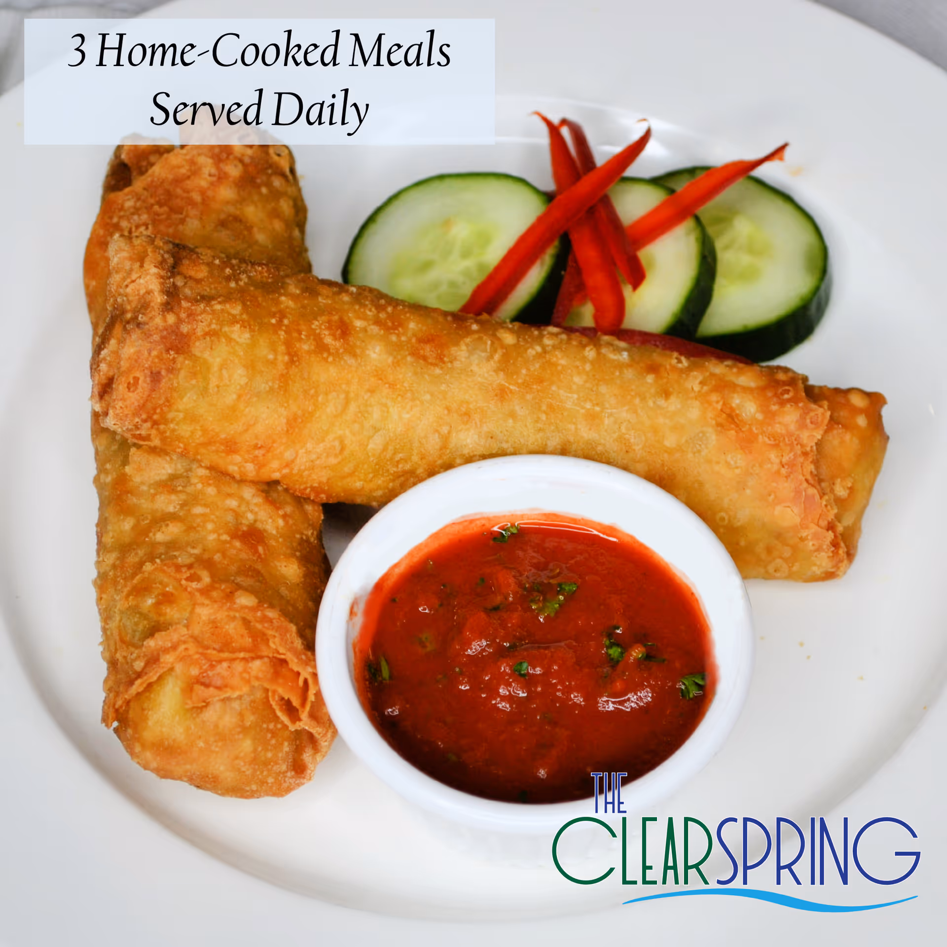 Two crispy fried egg rolls served on a white plate with a small bowl of red dipping sauce garnished with herbs, accompanied by cucumber slices and thin red pepper strips. Text on the image reads '3 Home-Cooked Meals Served Daily'.