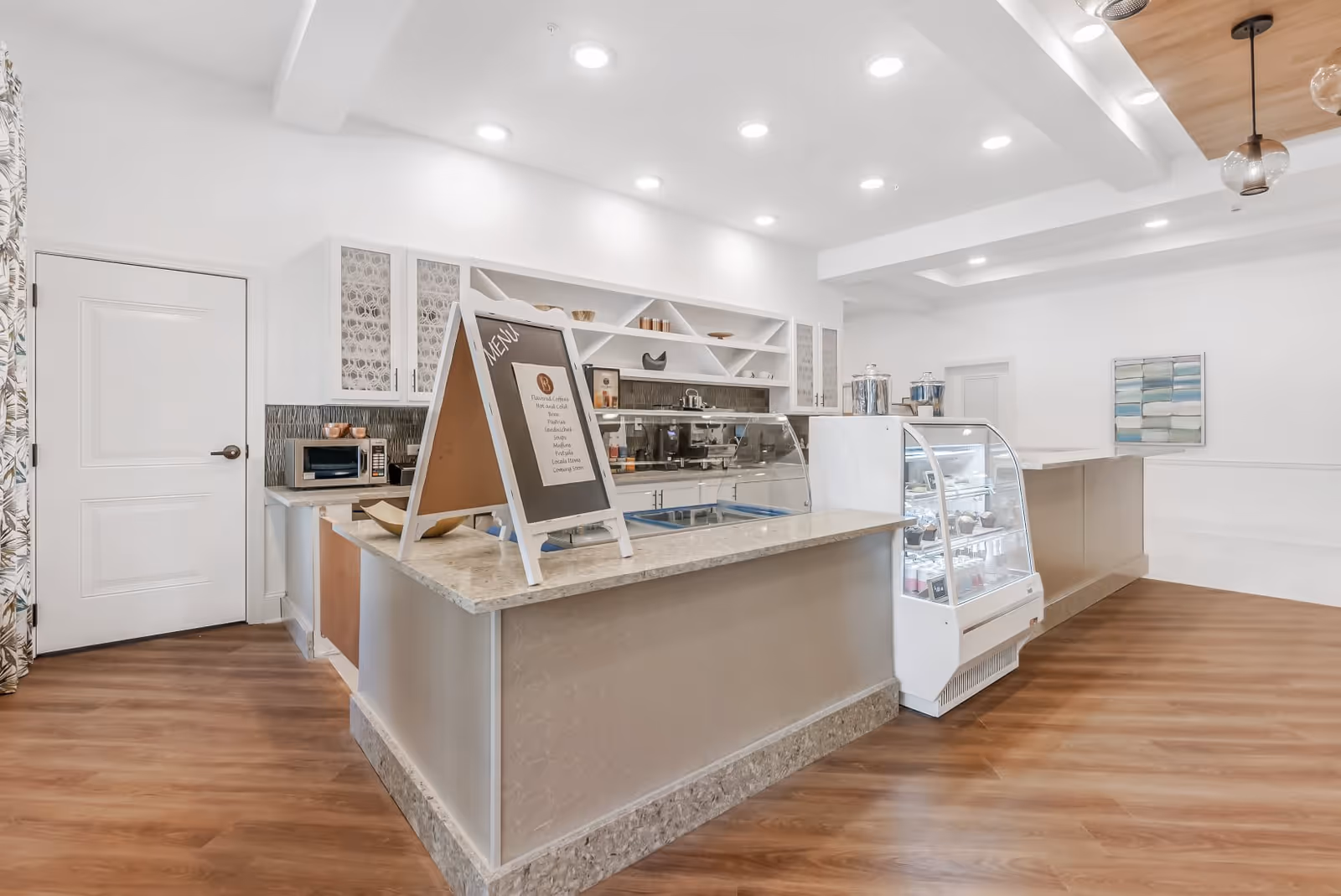 Bright and modern cafe counter area with a menu board on the counter, glass display case with desserts, coffee machine, microwave, and white cabinetry with decorative glass doors. The space has wood flooring, recessed lighting, and a piece of abstract wall art.