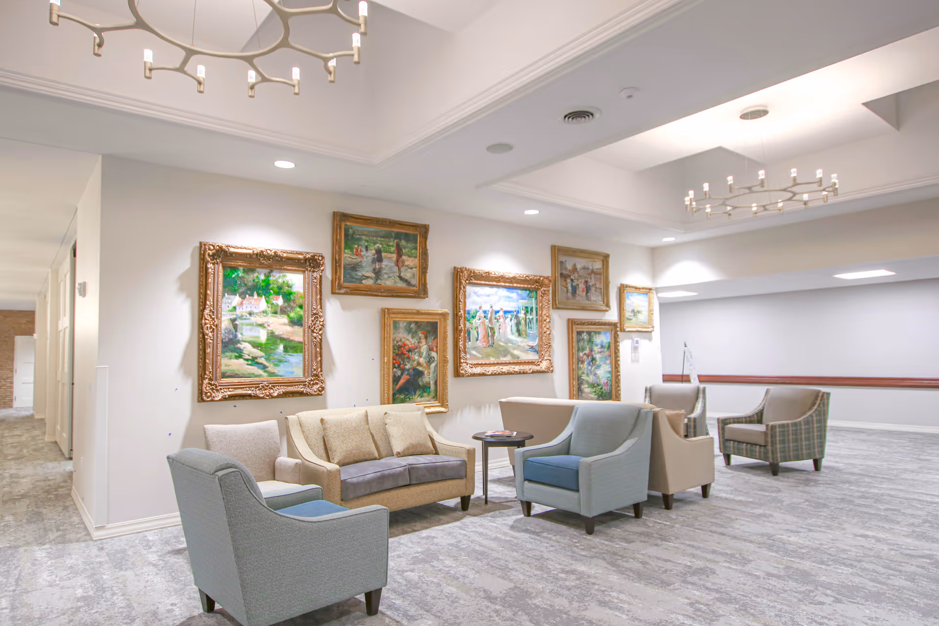 Spacious senior living common area with sofas and armchairs arranged around small tables beneath framed paintings.