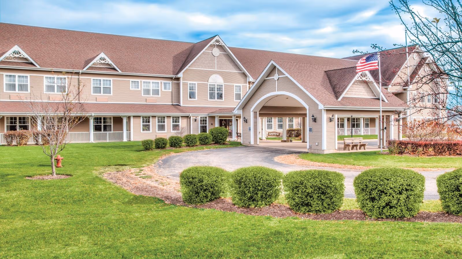 Exterior view of a two-story senior living facility building with beige siding and a red roof. The building has multiple windows, a covered entrance with a circular driveway, an American flag on a flagpole, green bushes, a small tree, and a well-maintained lawn under a partly cloudy sky.