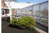Outdoor garden area with a white lattice fence, a small green bush, a bird feeder, and a paved walkway beside the fence. The area is part of a facility with a building visible in the background under a clear sky.