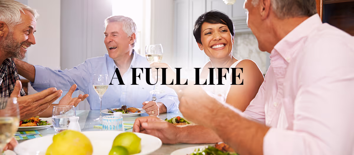 Four older adults sitting around a dining table, enjoying a meal and drinks while smiling and engaging in conversation. The table is set with plates of food, glasses of white wine, and water glasses. The text 'A FULL LIFE' is overlaid in the center of the image.