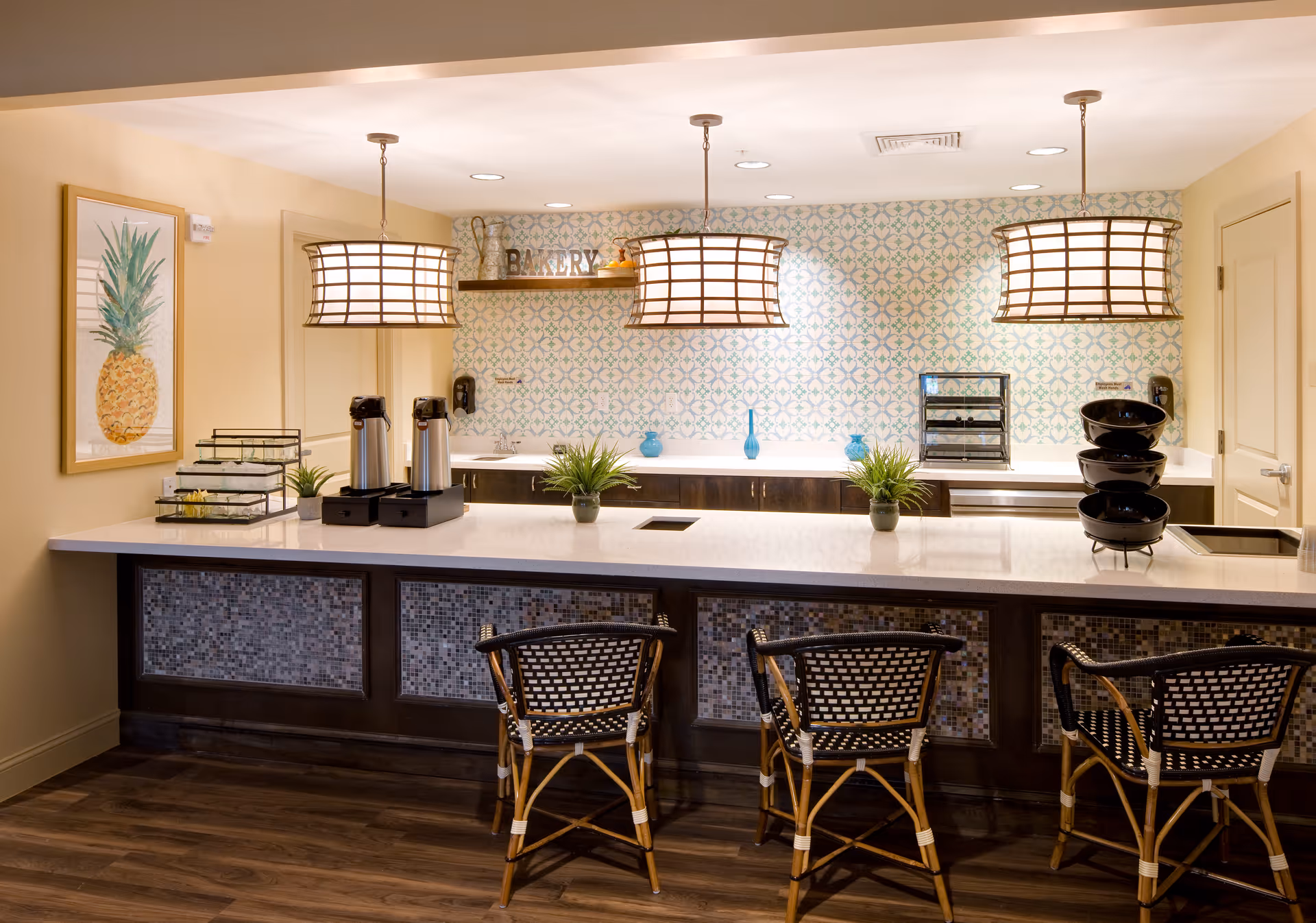 A bright and inviting kitchen or serving area with a long white countertop and three wicker chairs with black and white woven seats. Above the counter hang three large pendant lights with a grid pattern. The back wall features a decorative tile backsplash with a blue and green pattern, a shelf with the word 'BAKERY' displayed, and various small plants and decorative items. On the counter are two coffee dispensers, a tiered tray, and a stack of black bowls. The floor is wood, and a framed pineapple artwork hangs on the left wall.