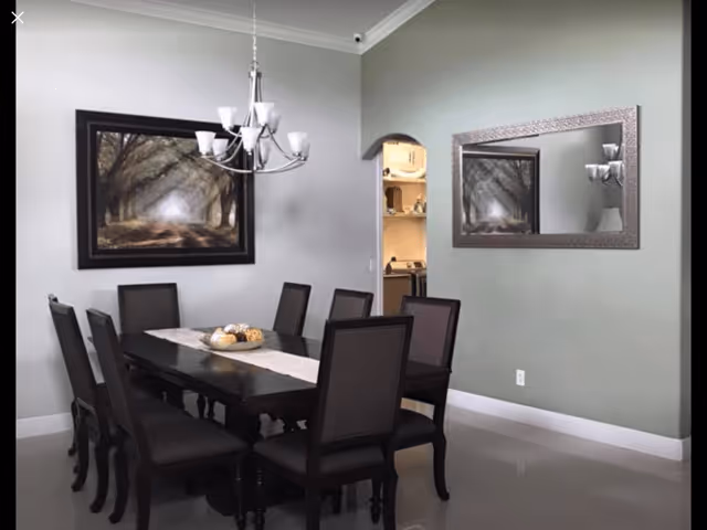 A dining room with a dark wooden rectangular table surrounded by eight matching chairs with dark upholstery. A chandelier with multiple lights hangs above the table. On the table is a decorative centerpiece with assorted items. The walls are painted light gray, featuring a large framed painting of a tree-lined path on one wall and a large rectangular mirror on the adjacent wall reflecting the painting. An open doorway leads to a kitchen area with visible shelves and appliances.