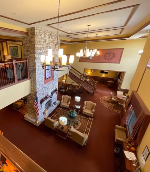 Interior view of a senior living facility lounge area with a large stone fireplace, several armchairs arranged around a coffee table, chandeliers hanging from the ceiling, and a staircase leading to an upper level. The room has warm yellow walls, red carpeting, and decorative wall art.