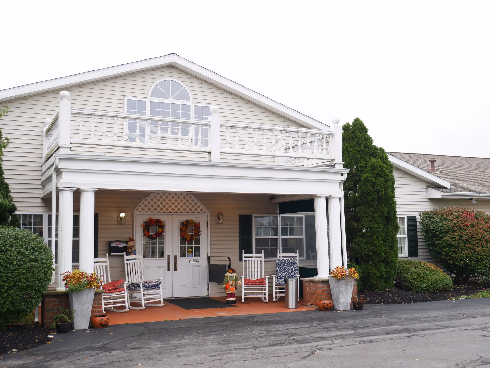 Front exterior view of a senior living facility named Seneca Lake Terrace, featuring a covered entrance with white columns, rocking chairs with cushions, autumn wreaths on double doors, and decorative plants on either side of the entrance.