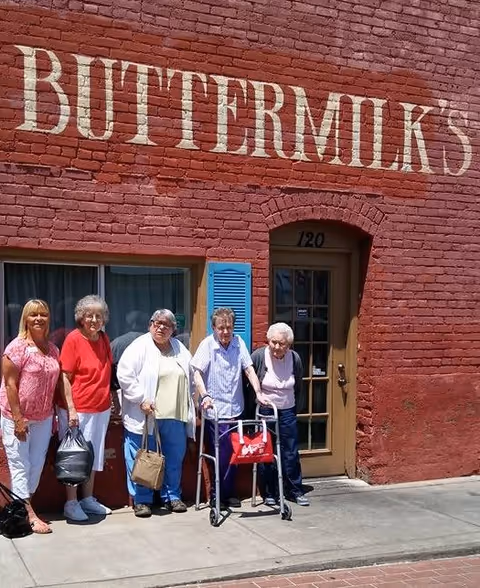 Five older women, two using walkers, stand on a sidewalk in front of a red brick building with a large painted sign reading "BUTTERMILK'S" above the door.