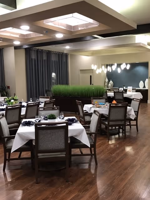 A dining room in a senior living facility with several tables covered in white tablecloths, each set with plates, glasses, and napkins. The room has wooden flooring, modern ceiling lights, and a decorative planter with green grass in the center. There are dark curtains on the windows and a sideboard with decorative vases and a modern light fixture on the back wall.