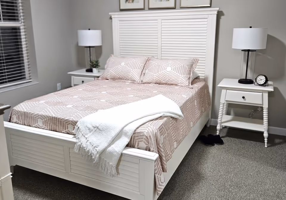 A neatly made bed with a white wooden frame and headboard, covered with a patterned beige and white bedspread and two matching pillows. A white throw blanket is draped over the foot of the bed. On each side of the bed are white nightstands with drawers, each holding a black-based lamp with a white shade. One nightstand also has a small clock and the other has a small plant. The room has gray carpet and a window with white blinds on the left wall.