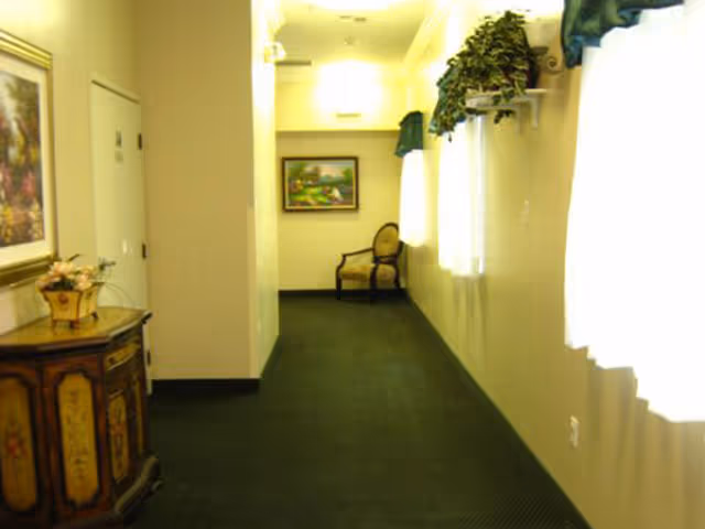 A carpeted hallway in a senior living facility with a decorative cabinet, framed paintings, and a chair at the far end.