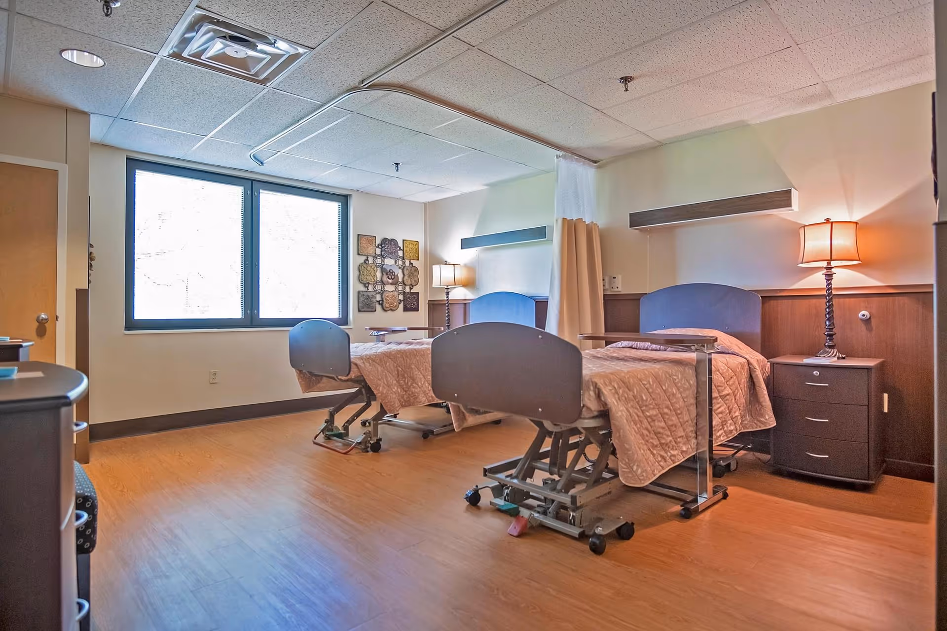 A shared resident bedroom with two adjustable hospital-style beds, bedside tables, a lamp, and a large window.
