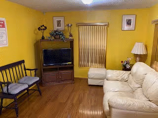 A cozy living room with yellow walls and wooden flooring. The room features a beige leather sofa, a wooden bench with cushions, a wooden TV stand with a flat-screen television, and a small ottoman. There are framed pictures on the walls, a table lamp on a side table, and a window with vertical blinds.