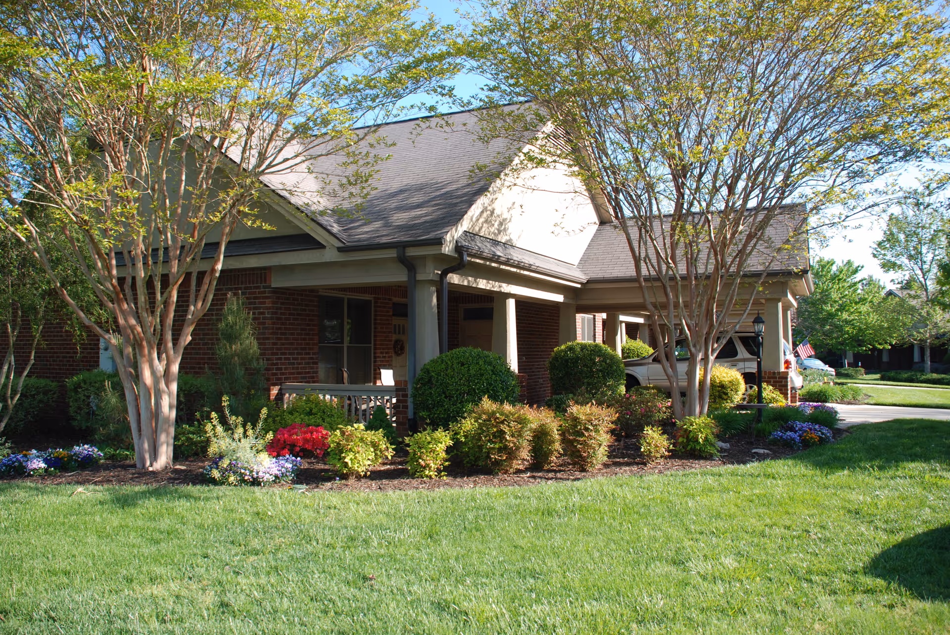 Exterior view of a brick building with a covered porch surrounded by well-maintained landscaping including bushes, flowering plants, and trees under a clear blue sky.