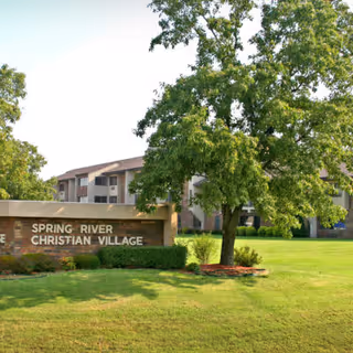 Exterior view of a senior living facility with a large green lawn, a tree, and a sign that reads 'Spring River Christian Village'. The building is visible in the background under a clear sky.
