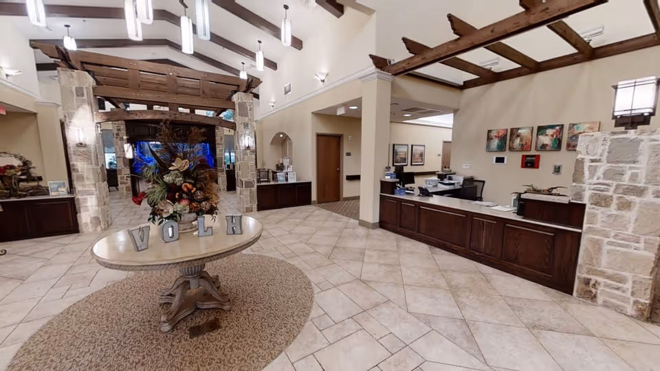Spacious reception area with a round table in the center displaying a floral arrangement and decorative letters spelling 'VOLH'. The room features tiled flooring, stone pillars, wooden beams on the ceiling, and a reception desk with office equipment and colorful artwork on the wall behind it.