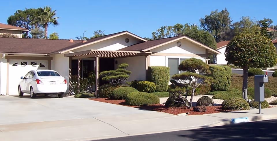 Front exterior of a single-story house with a driveway and white car, trimmed shrubs, and a mailbox.