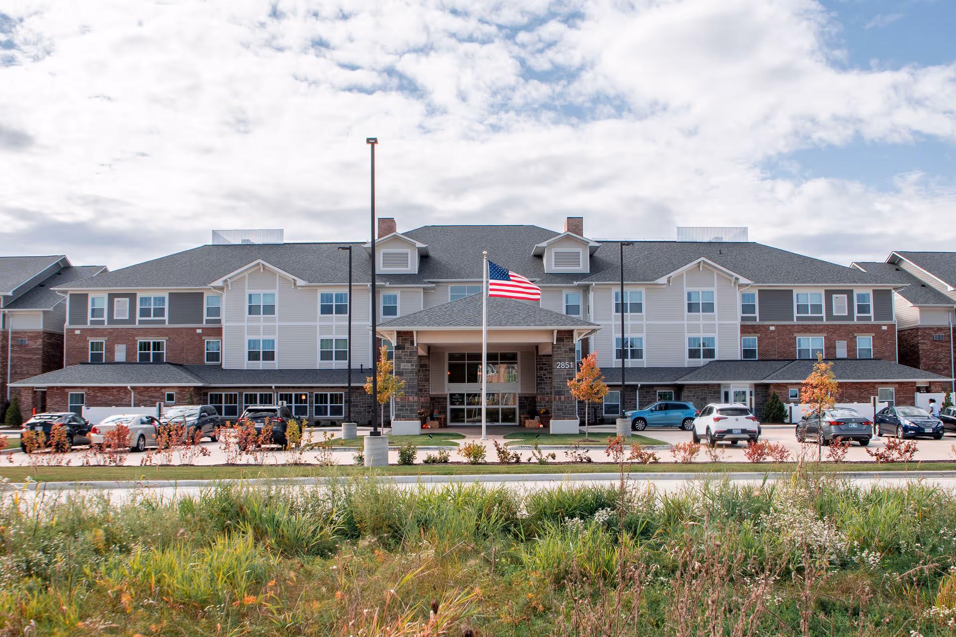 Front exterior view of Quarry Ridge Retirement Community building with an American flag on a flagpole in front, several cars parked along the driveway, and a grassy area with plants in the foreground under a partly cloudy sky.