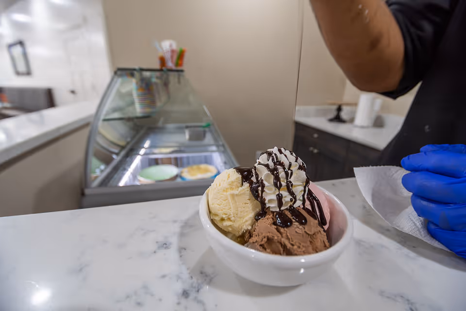A bowl of ice cream topped with whipped cream and chocolate syrup sits on a marble counter beside a server wearing blue gloves and an ice cream display case in the background.