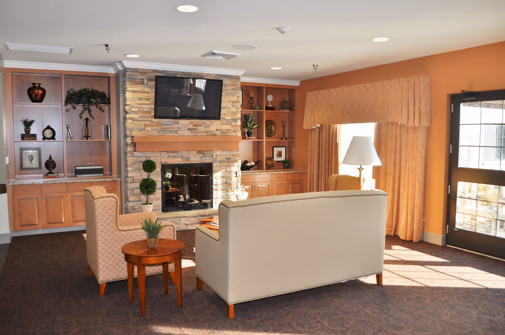 A cozy living room area with a beige sofa and two patterned armchairs arranged around a stone fireplace with a mounted flat-screen TV above it. The room features built-in wooden shelves with decorative items, a small round wooden table with a plant, a floor lamp, and large windows with orange curtains letting in natural light.