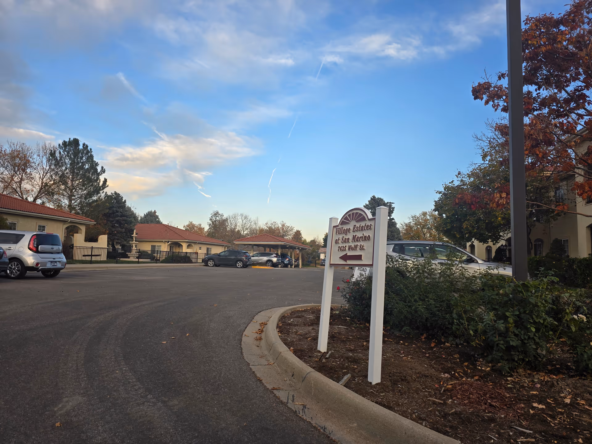 Parking lot area of San Marino Retirement Community with several parked cars, a sign pointing to Village Estates of San Marino at 7435 Wolff St., surrounded by trees with autumn foliage and buildings with red-tiled roofs under a partly cloudy blue sky.
