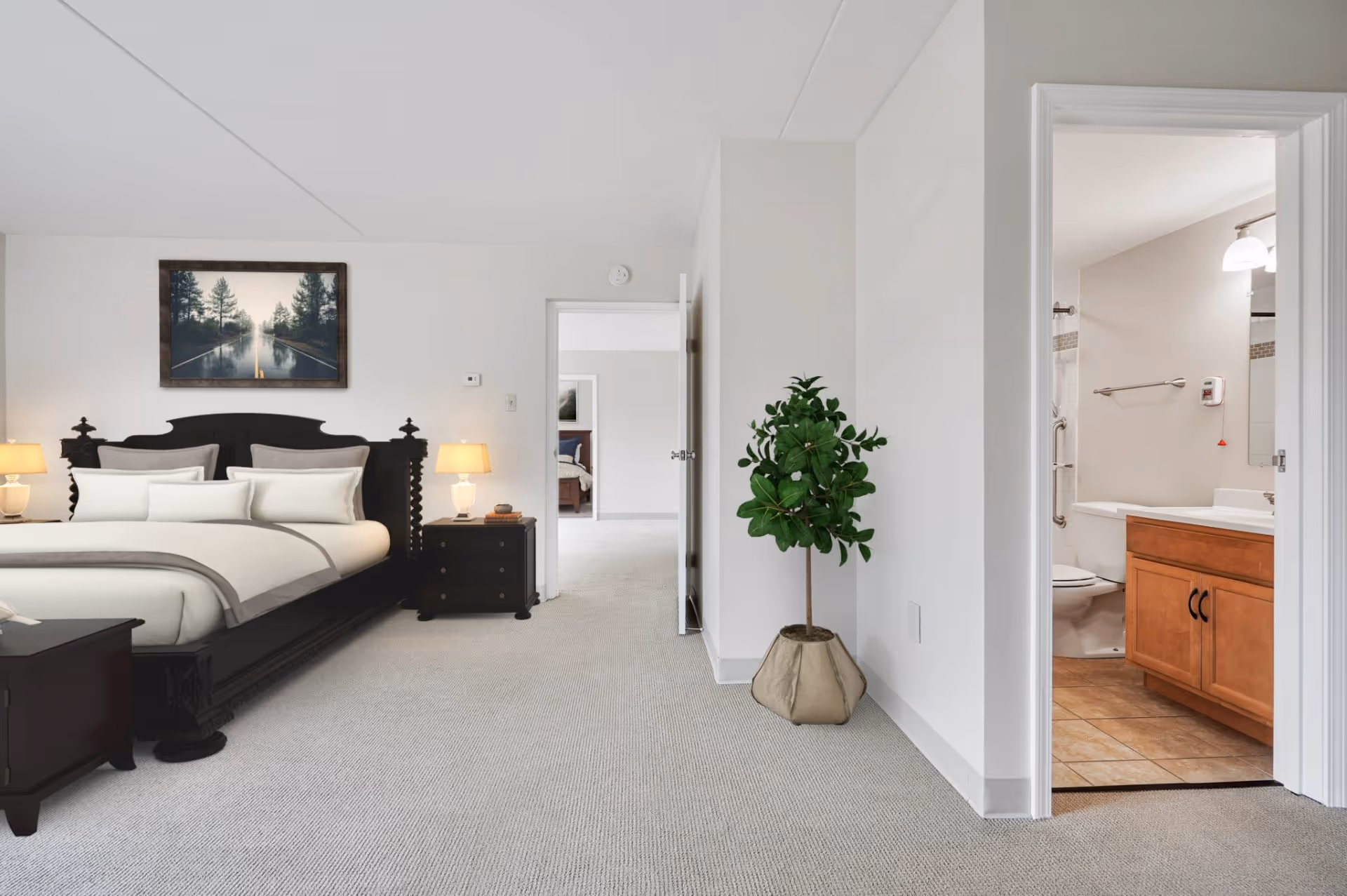 A bright bedroom with a large dark wooden bed with white and gray bedding, two bedside tables with lamps, a framed picture of a forest and road above the bed, a potted plant in a beige fabric planter on the floor, and an open door leading to a bathroom with a wooden vanity, toilet, and tiled floor.