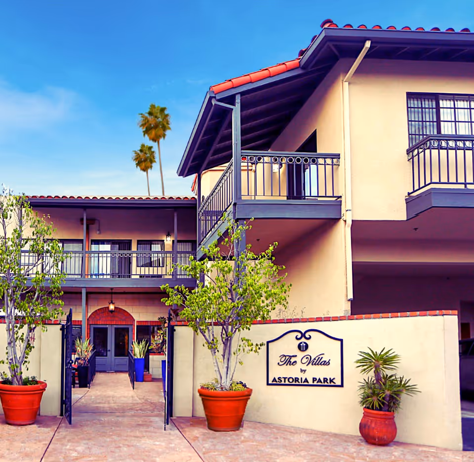 Exterior view of The Villas by Astoria Park, showing a two-story building with balconies, potted plants, and an entrance gate under a clear blue sky.