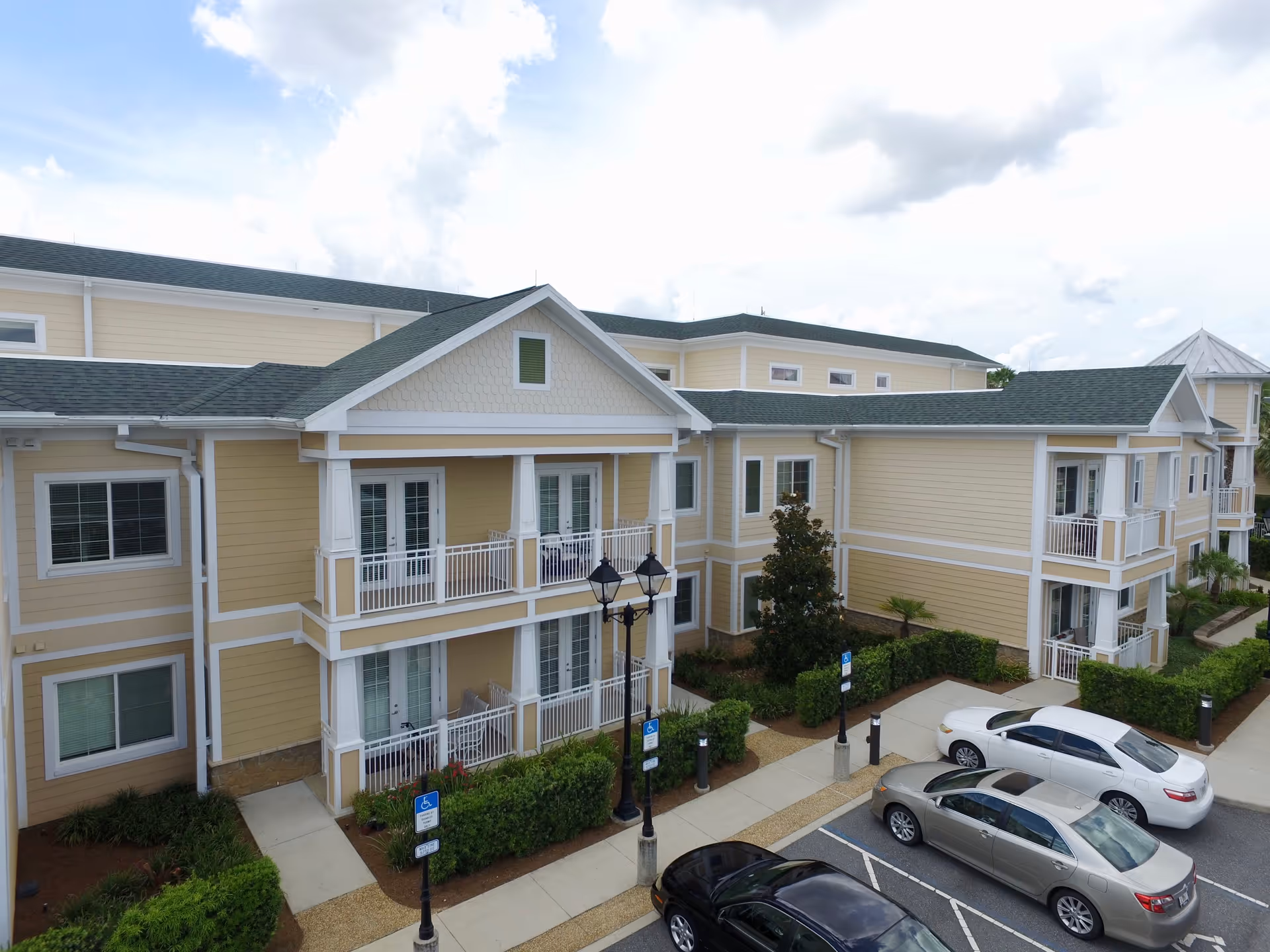 Exterior view of a two-story senior living facility building with beige siding and white trim. The building features balconies with white railings, several windows, and a green roof. There are three cars parked in designated parking spaces in front of the building, along with landscaped bushes and a sidewalk. The sky is partly cloudy.